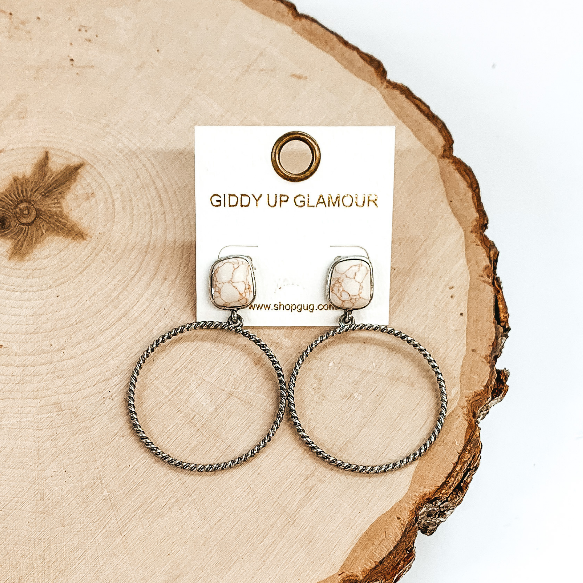 Rounded, square stone stud earrings in ivory with a silver, twisted open circle pendant hanging from the bottom. These earrings are pictured on a piece of wood on a white background.