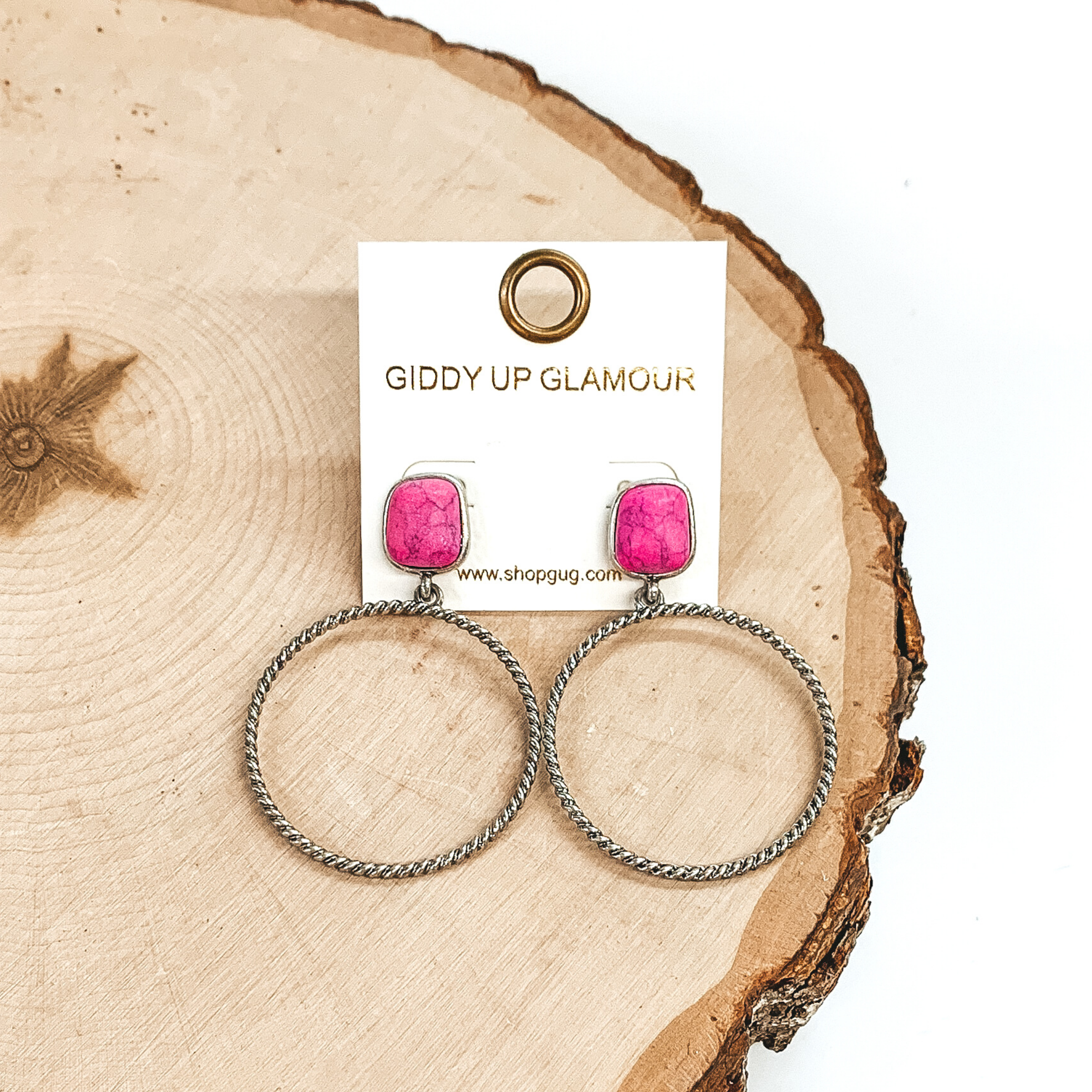 Rounded, square stone stud earrings in pink with a silver, twisted open circle pendant hanging from the bottom. These earrings are pictured on a piece of wood on a white background. 