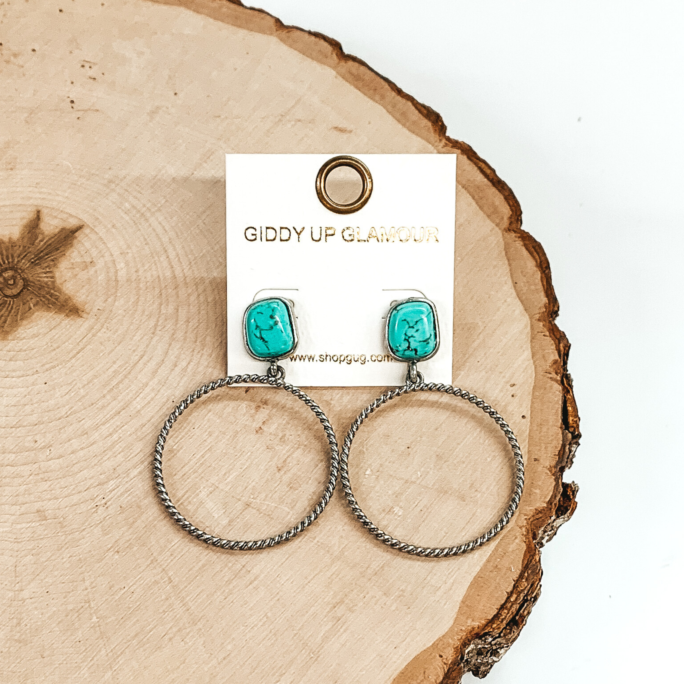 Rounded, square stone stud earrings in turquoise with a silver, twisted open circle pendant hanging from the bottom. These earrings are pictured on a piece of wood on a white background. 
