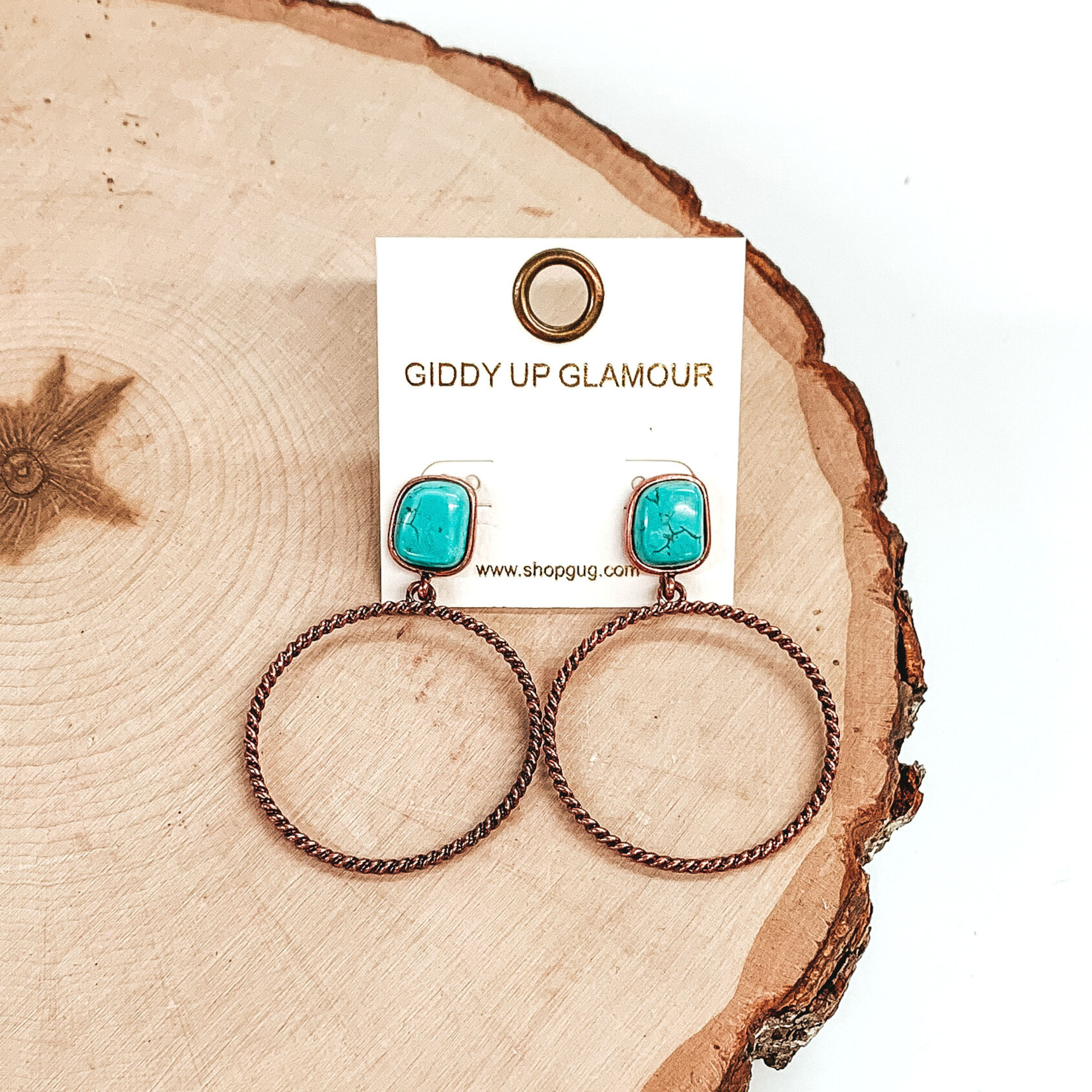 Rounded, square stone stud earrings in turquoise with a copper, twisted open circle pendant hanging from the bottom. These earrings are pictured on a piece of wood on a white background. 