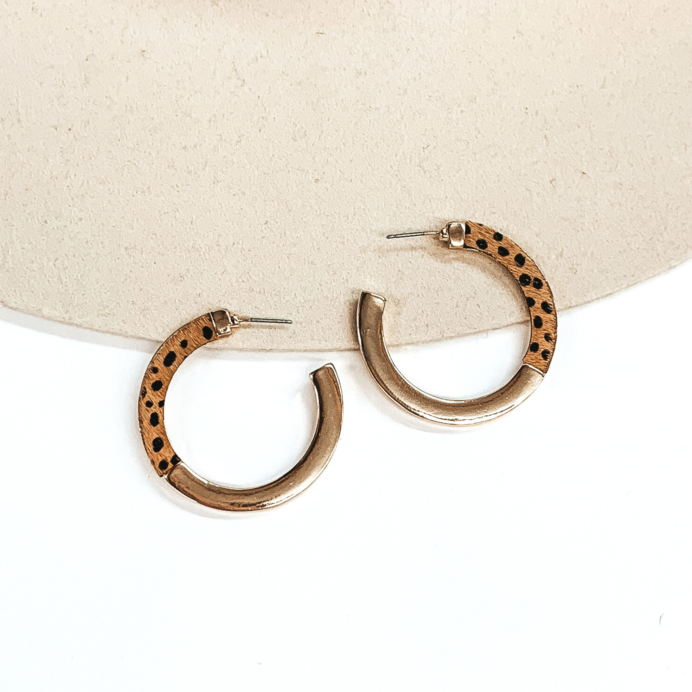 Flat hoops that are half gold and half tan with a black cheetah print. These earrings are pictured on a white and beige background.