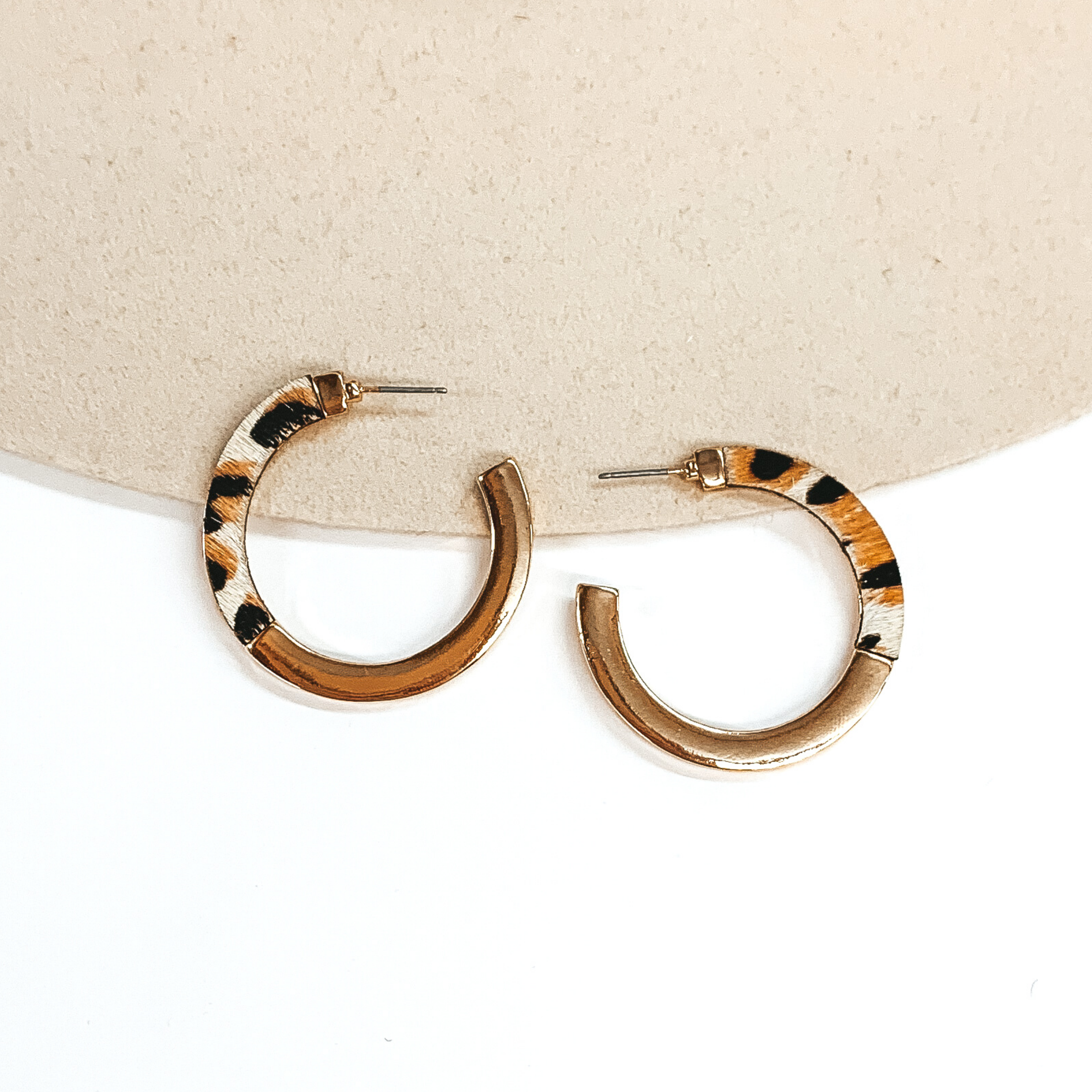 Flat hoops that are half gold and half ivory with a black and brown leopard print. These earrings are pictured on a white and beige background. 