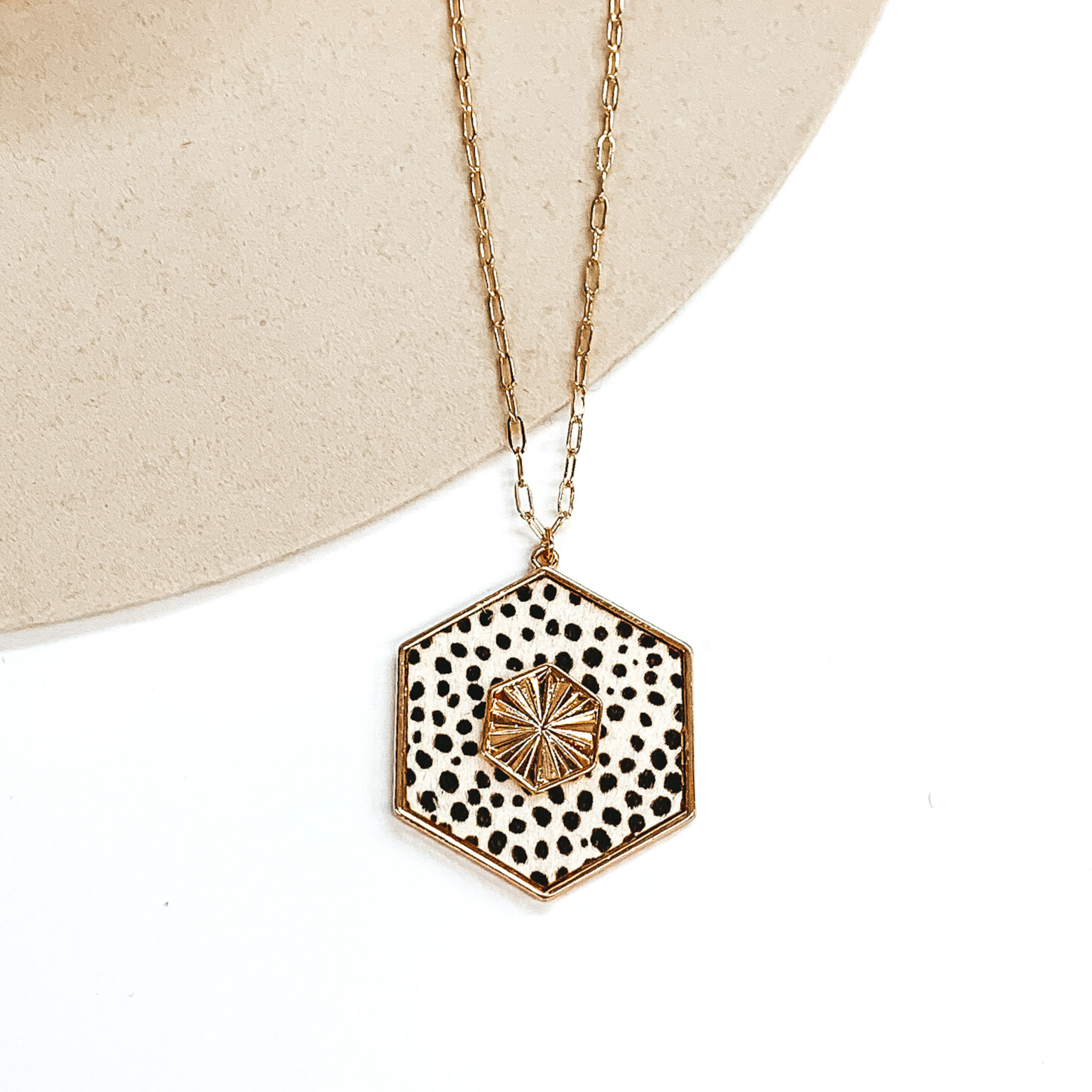 Thin, gold chained necklace with an ivory hexagon pendant with a black cheetah print. The pendant is outlined in gold and has a gold, textured hexagon charm in the center. This necklace is pictured on a white and beige background. 