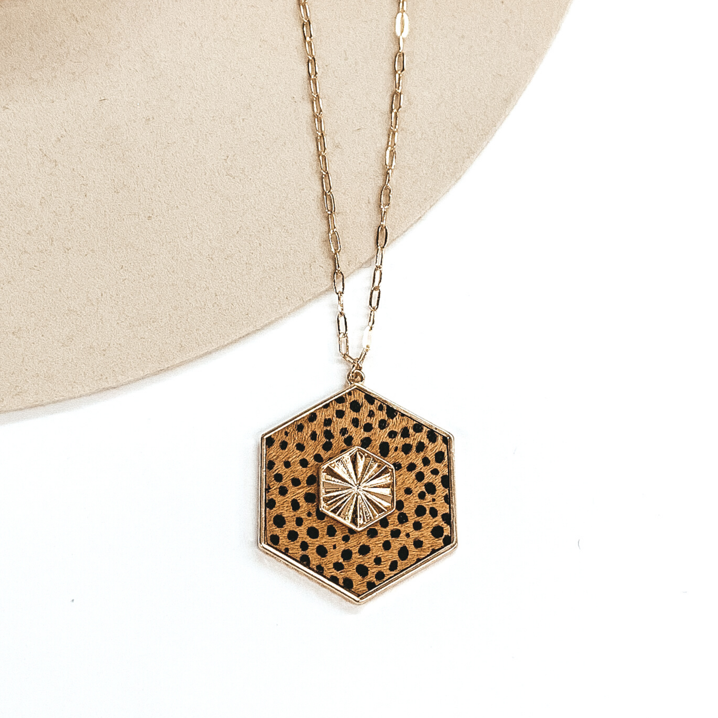 Thin, gold chained necklace with an tan hexagon pendant with a black cheetah print. The pendant is outlined in gold and has a gold, textured hexagon charm in the center. This necklace is pictured on a white and beige background.
