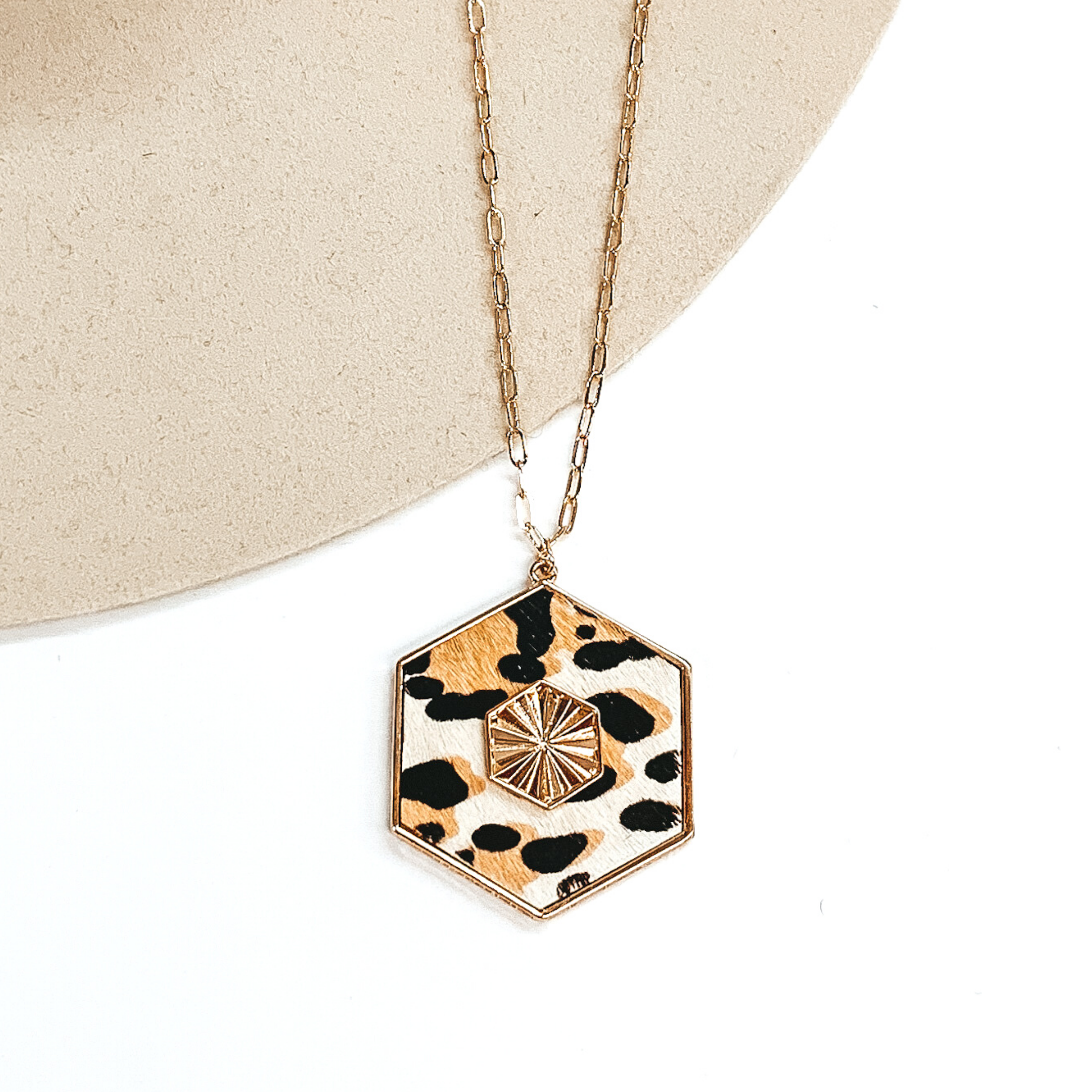 Thin, gold chained necklace with an ivory hexagon pendant with a black and tan leopard print. The pendant is outlined in gold and has a gold, textured hexagon charm in the center. This necklace is pictured on a white and beige background. 