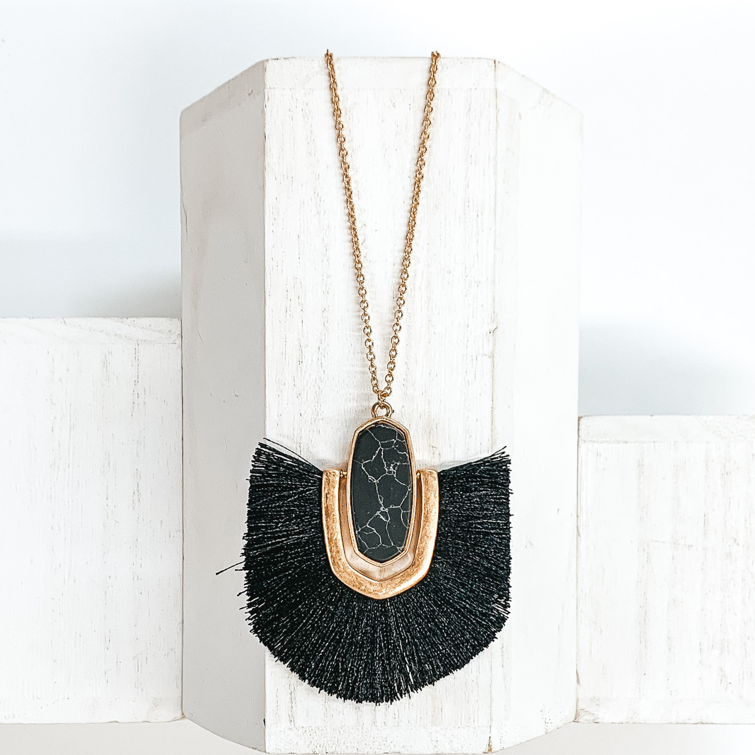 Thin, gold chained necklace with an oval shaped black stone pendant that has a half oval gold accent connected at the sides that inlcudes black fringe. This necklace is pictured on a white block on a white background.