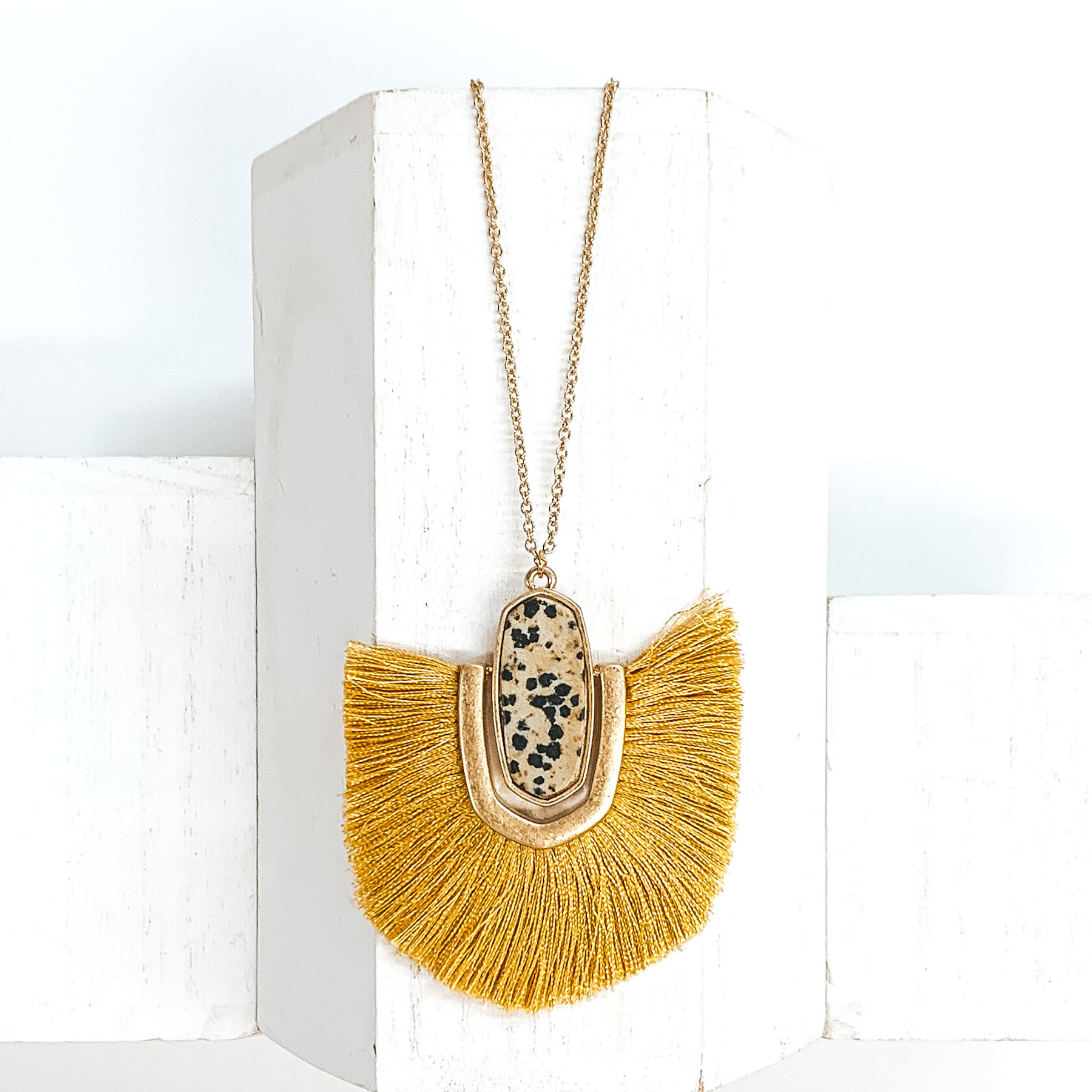 Thin, gold chained necklace with an oval shaped beige stone with black dots pendant that has a half oval gold accent connected at the sides that inlcudes yellow fringe. This necklace is pictured on a white block on a white background. 