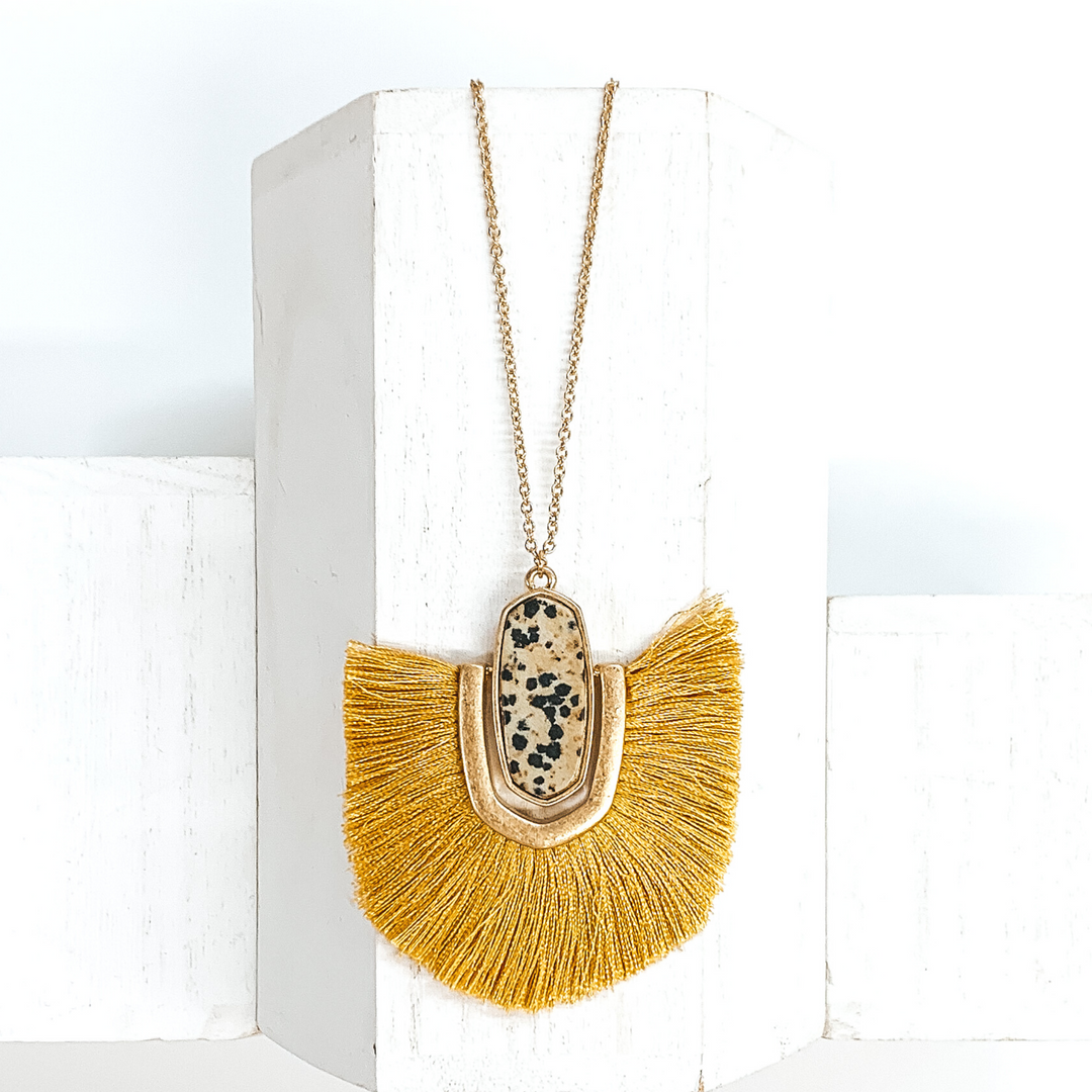 Thin, gold chained necklace with an oval shaped beige stone with black dots pendant that has a half oval gold accent connected at the sides that inlcudes yellow fringe. This necklace is pictured on a white block on a white background. 