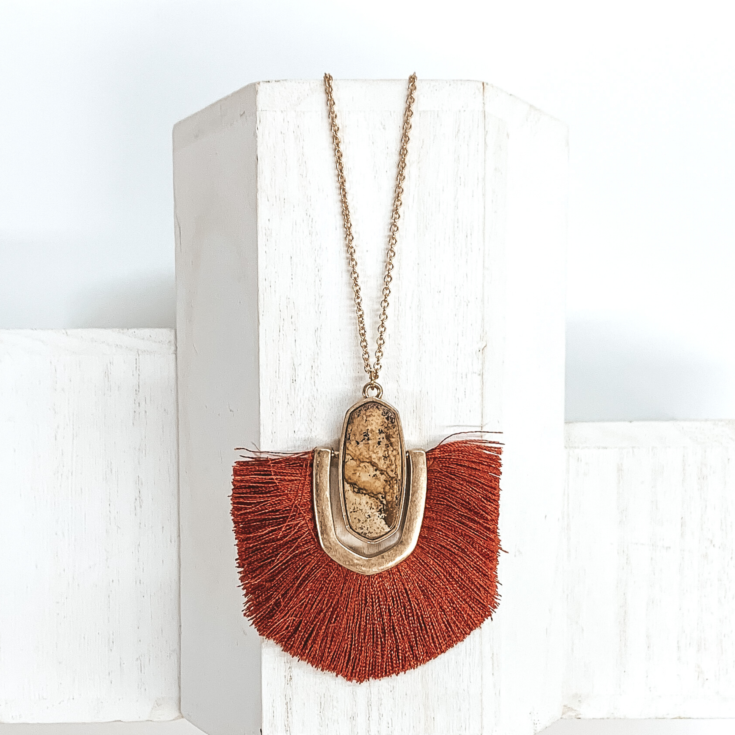 Thin, gold chained necklace with an oval shaped brown stone pendant that has a half oval gold accent connected at the sides that inlcudes rust red fringe. This necklace is pictured on a white block on a white background. 