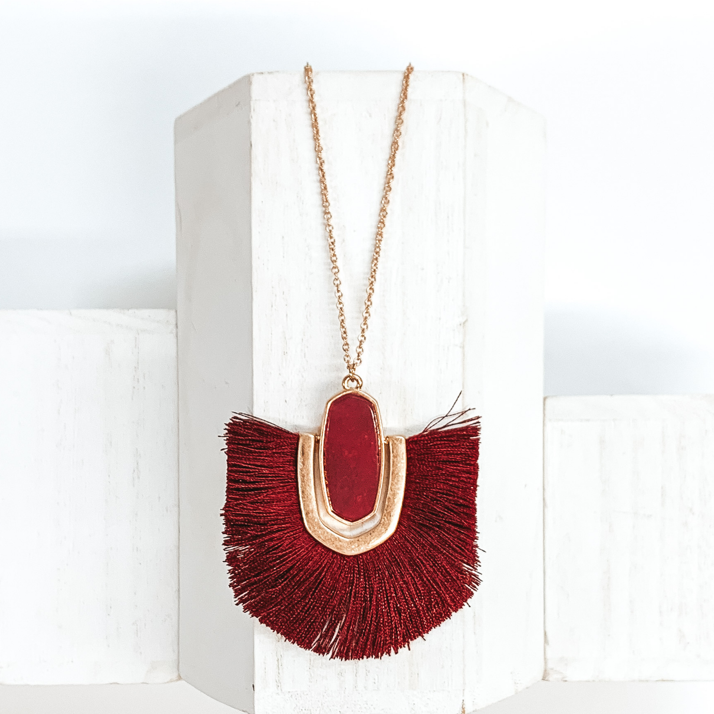 Thin, gold chained necklace with an oval shaped red stone pendant that has a half oval gold accent connected at the sides that inlcudes dark red fringe. This necklace is pictured on a white block on a white background.