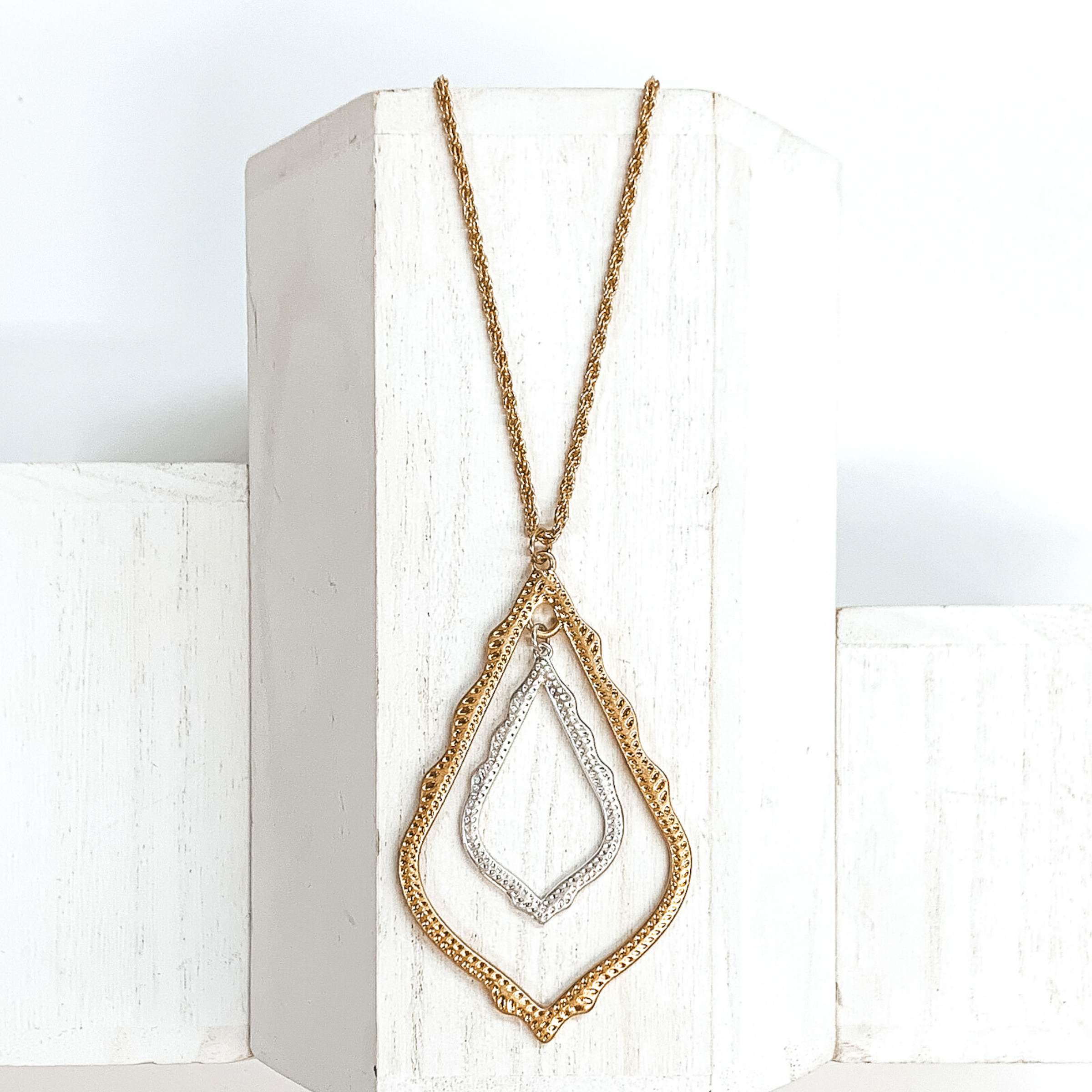 Small, gold rope chained necklace with two open teardrop pendants. This bigger pendant is gold and the smaller pendant is silver. This necklace is pictured on a white block on a white background.