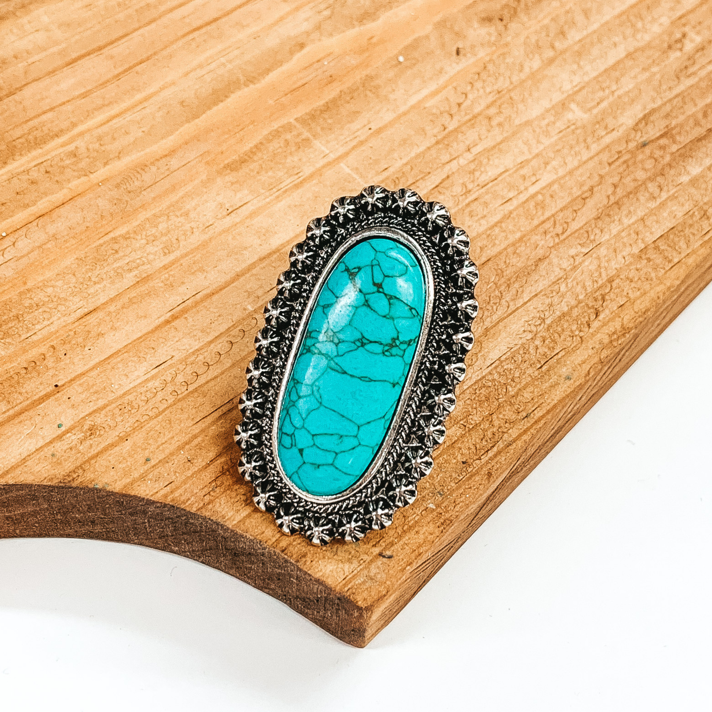 Silver cuff ring with oval, turquoise stone and silver, intricate detailed outline. This ring is pictured on a brown block on a white background.