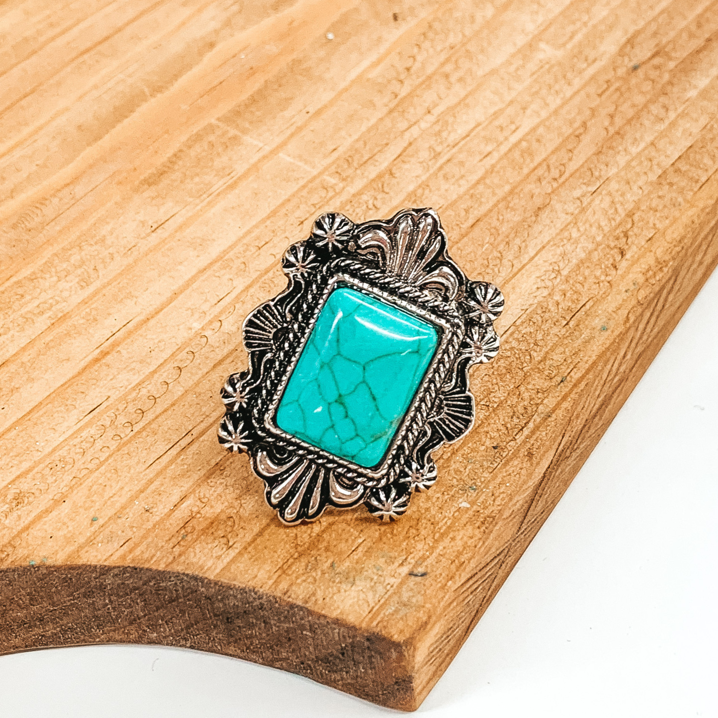 Silver cuff ring with reactangle, turquoise stone and silver, intricate detailed outline. This ring is pictured on a brown block on a white background. 