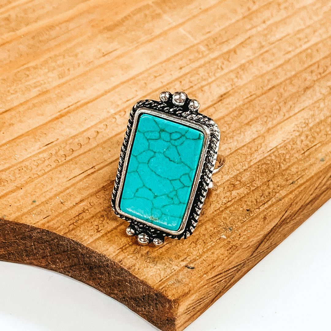 Silver cuff ring with reactangle, turquoise stone and silver detailed outline. This ring is pictured on a brown block on a white background. 