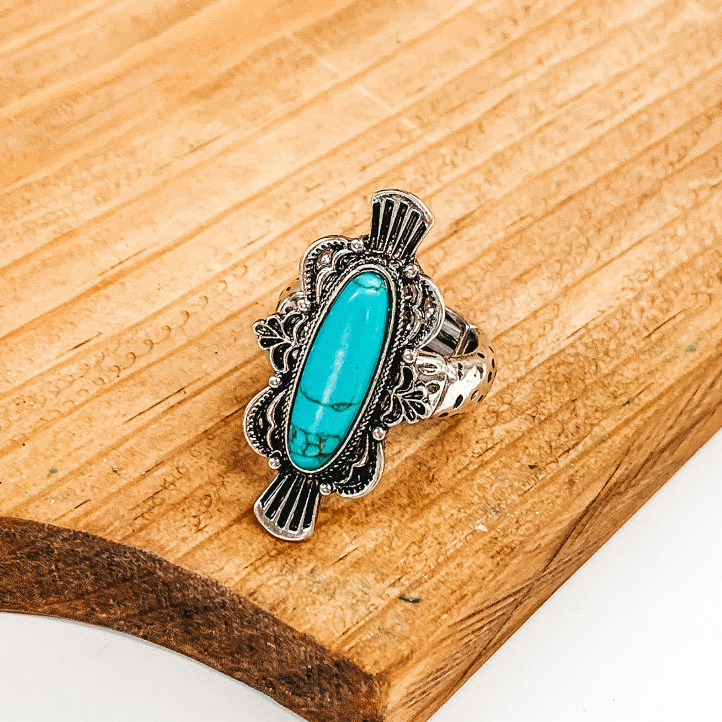 This ring is a silver stretchy ring with a thin band that has an intricate oval pendant with a center turquoise, oval stone. This ring is pictured laying on a brown block on a white background.