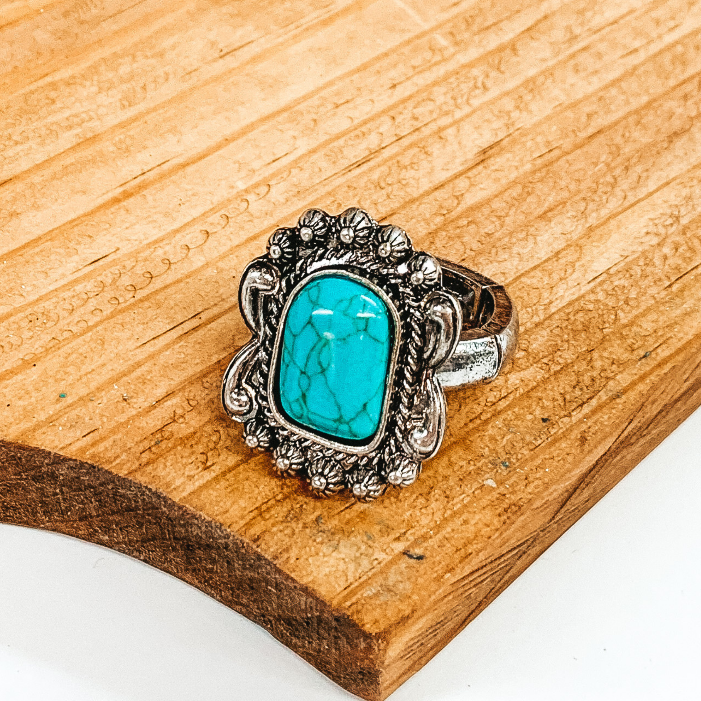 Silver stretchy ring that has a silver rectangular pendant with engraving and carving  detailing. In the center of the pendant, there is a turquoise rectangular stone. This ring is pictured laying on a brown block on a white background. 