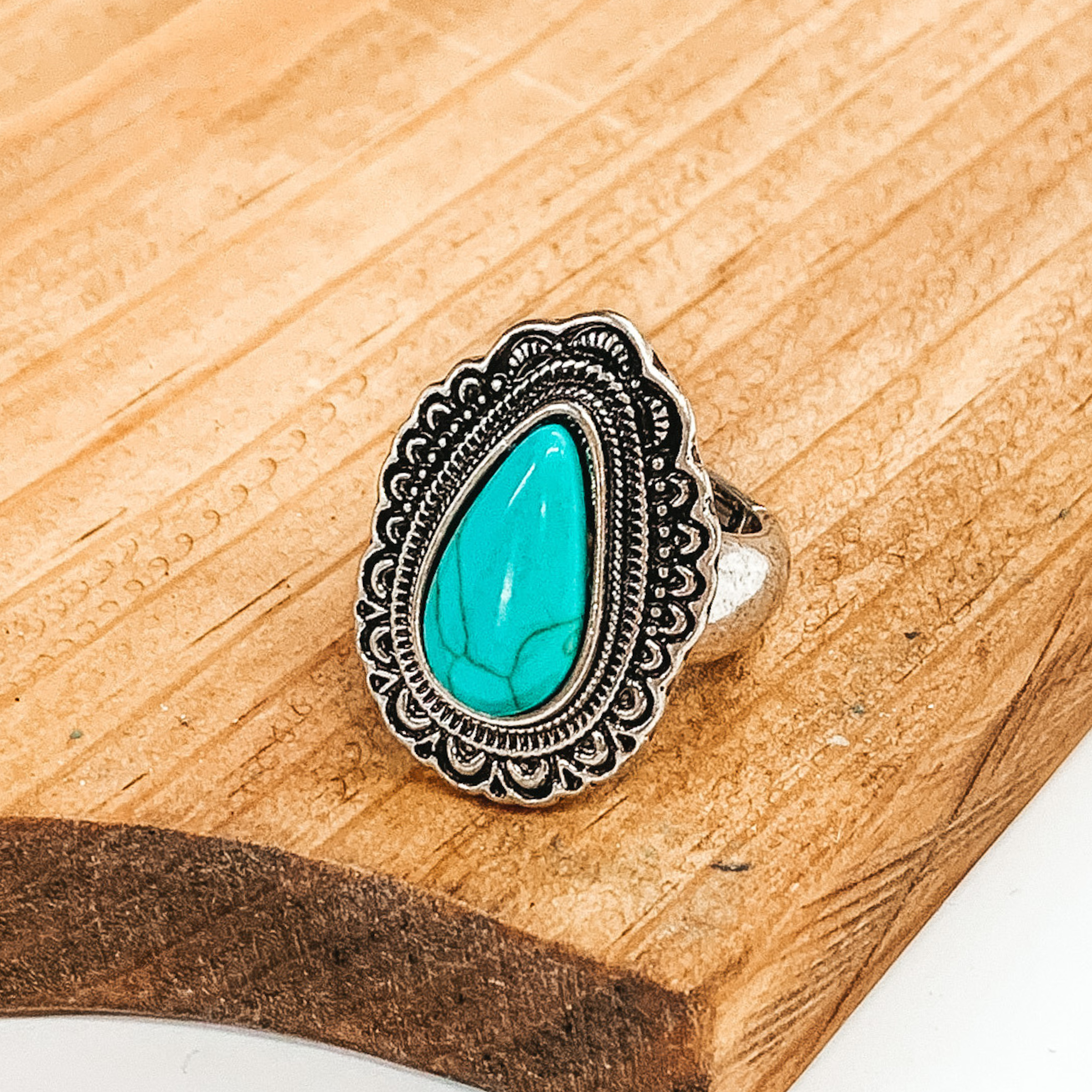 Silver stretchy ring that has a silver teardrop pendant with engraving detailing. In the center of the pendant, there is a turquoise teardrop stone. This ring is pictured laying on a brown block on a white background. 