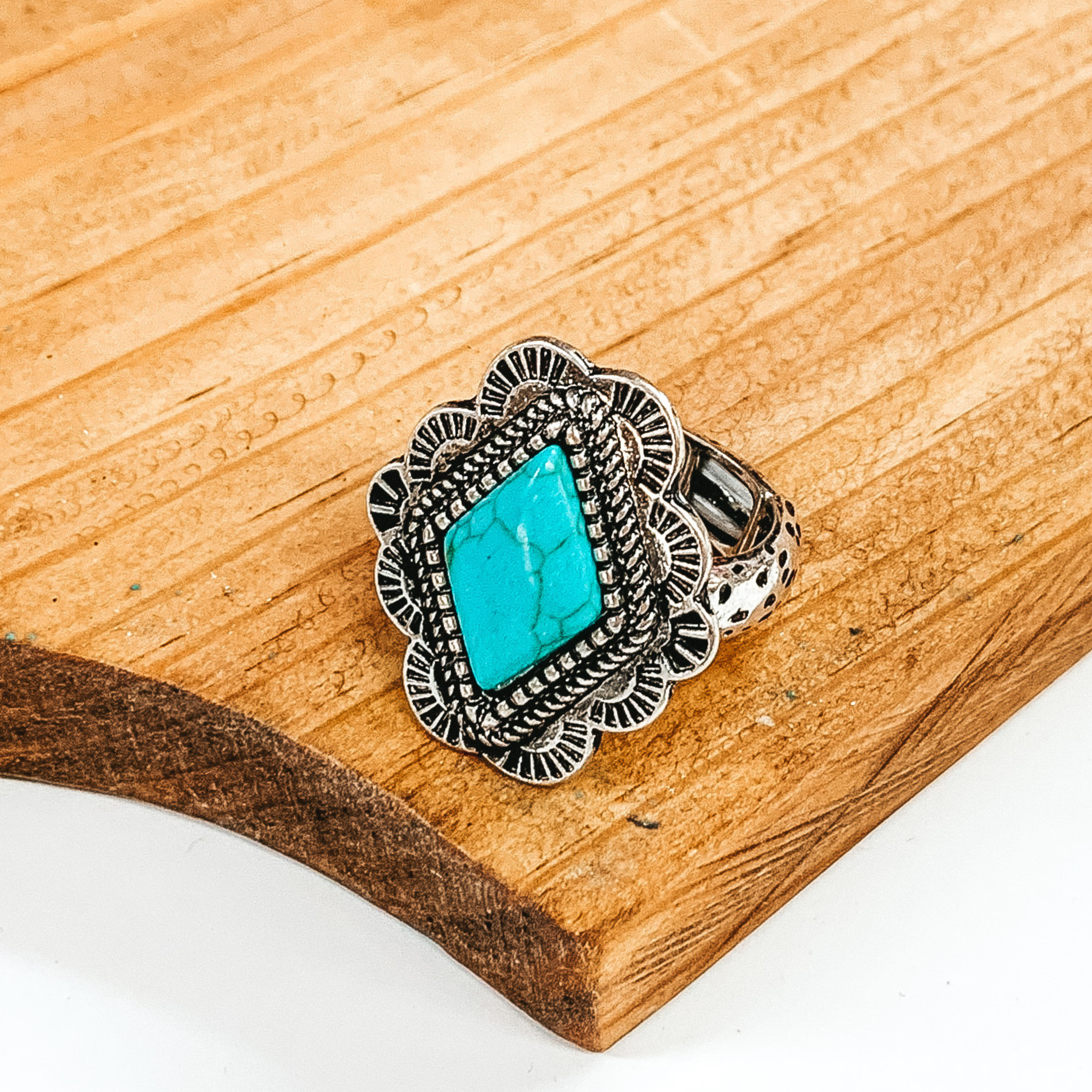 Silver stretchy ring that has a silver, rounded, diamond pendant with engraving detailing. In the center of the pendant, there is a turquoise diamond stone. This ring is pictured laying on a brown block on a white background.