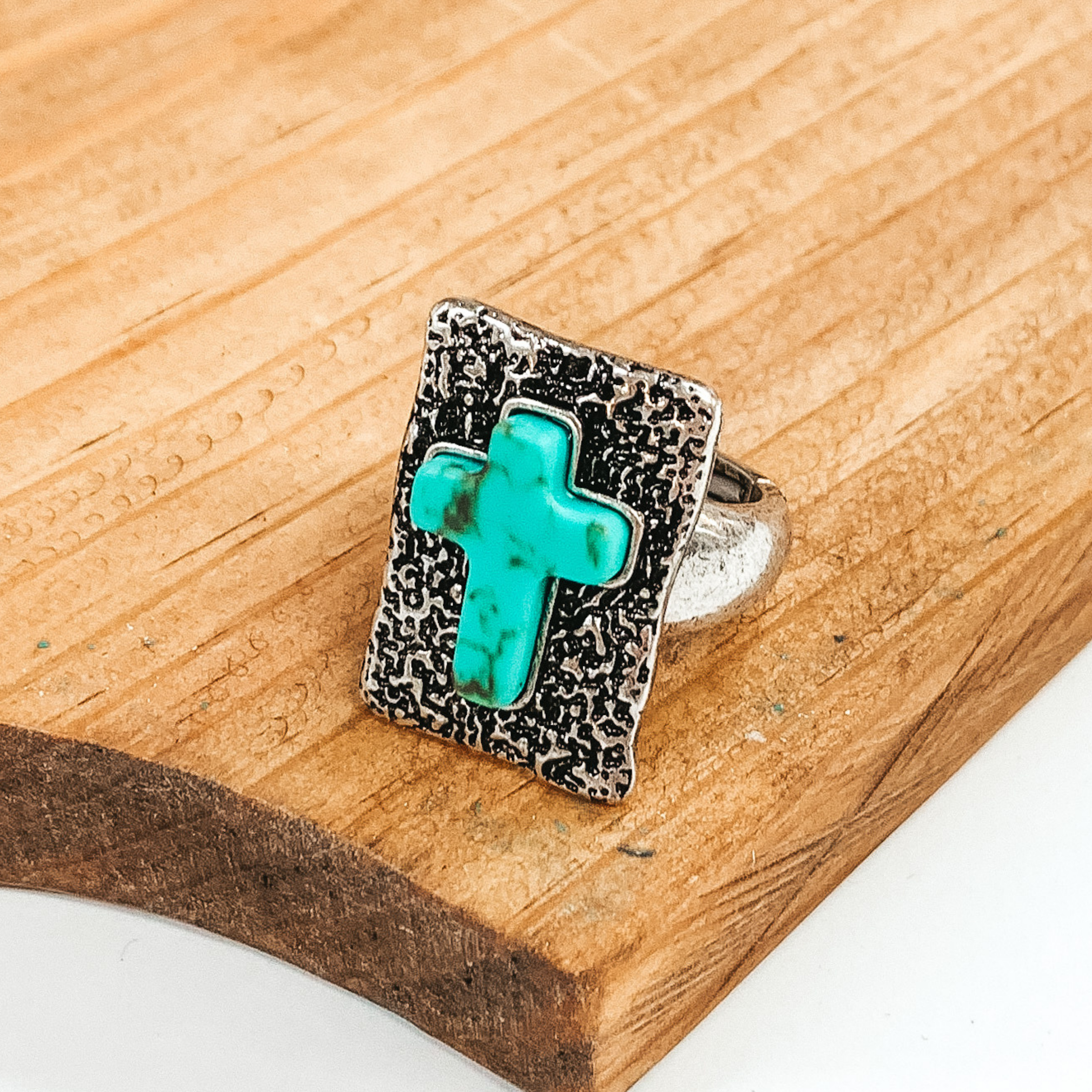 Silver stretchy ring that has a silver rectangle pendant with engraving detailing. In the center of the pendant, there is a turquoise cross stone. This ring is pictured laying on a brown block on a white background.