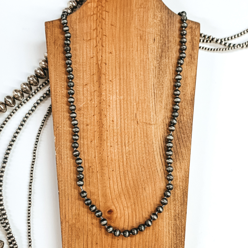 Silver beaded necklace that is pictured laying on a brown necklace holder. In the background there are silver beads and all is pictured on a white background.