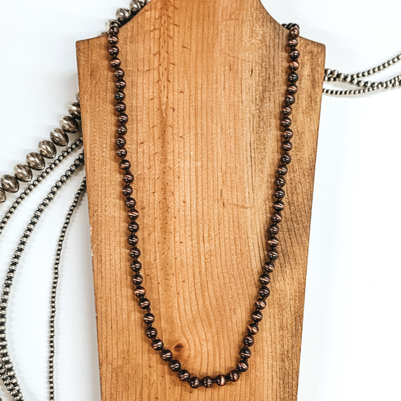 Copper beaded necklace that is pictured laying on a brown necklace holder. In the background there are silver beads and all is pictured on a white background.