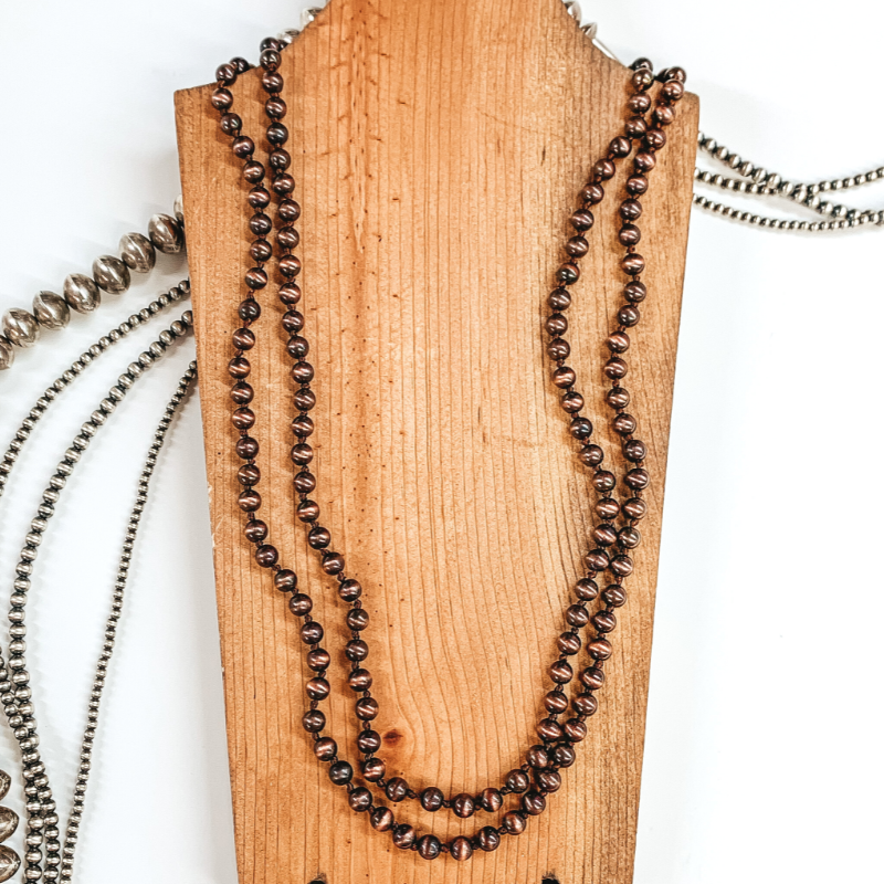 Copper beaded necklace that is picturede wrapped around twice on a brown necklace holder. In the background there are silver beads and all is pictured on a white background. 