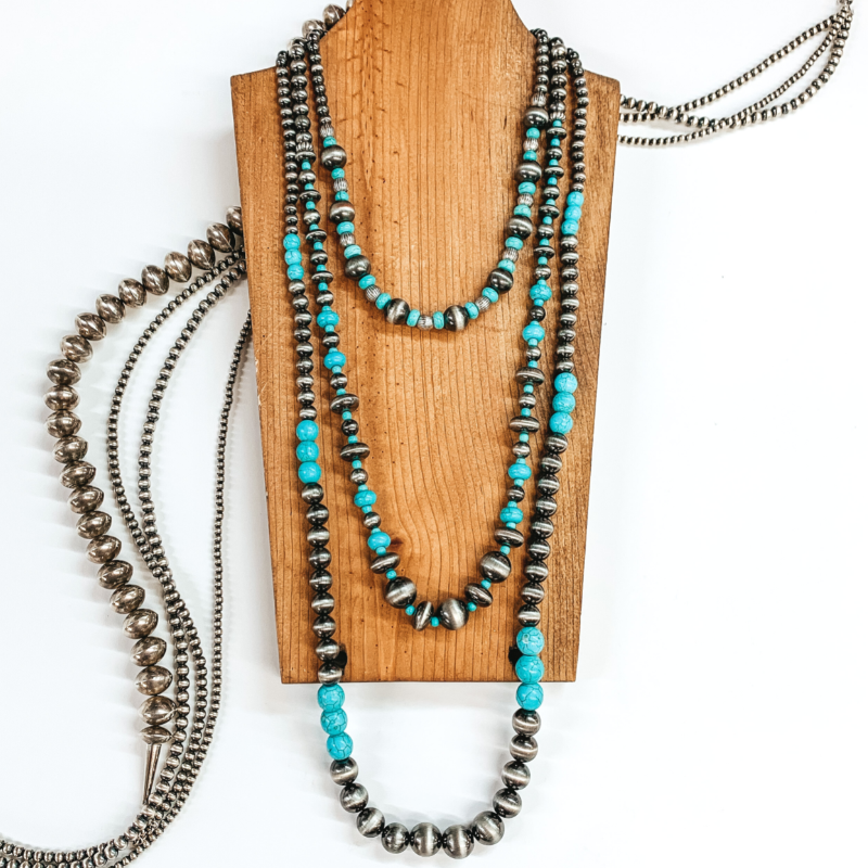 Three stranded beaded necklace. The longest strand has silver colored beads with turquoise beaded segments throughout. The medium length strand has silver beads that vary in size with turquoise beaded spacers. The shortest strand has silver beads and turquoise beads on after another. This necklace is pictured on a brown necklace holder on a white background with silver beads underneath the necklace holder.