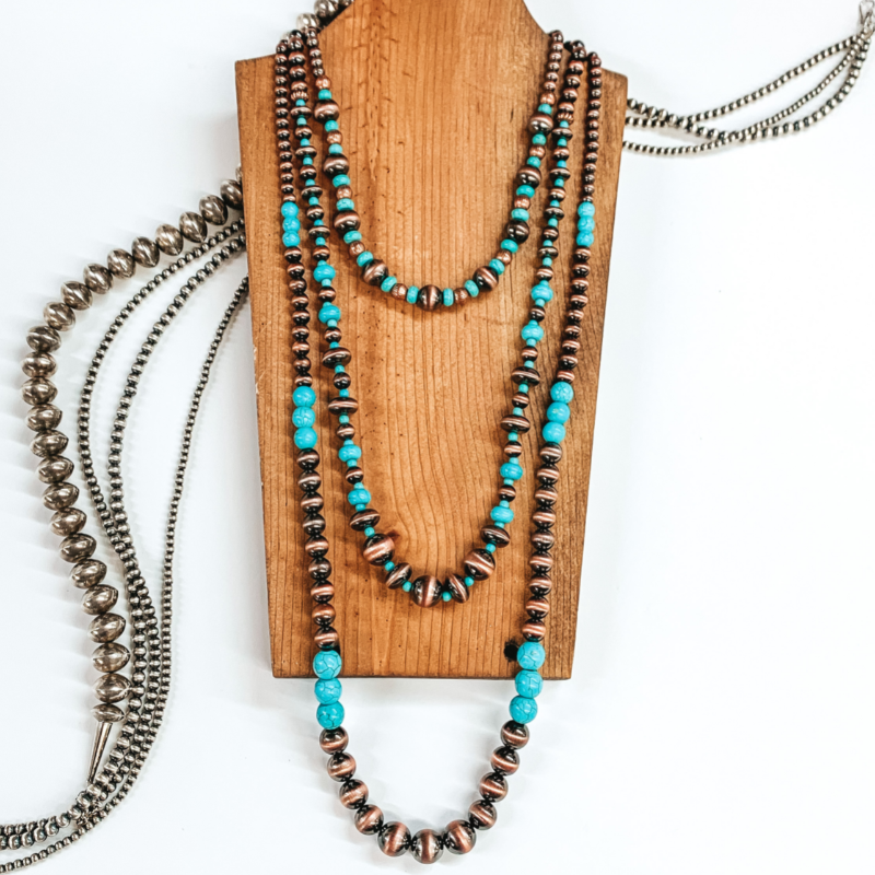 Three stranded beaded necklace. The longest strand has copper colored beads with turquoise beaded segments throughout. The medium length strand has copper beads that vary in size with turquoise beaded spacers. The shortest strand has copper beads and turquoise beads on after another. This necklace is pictured on a brown necklace holder on a white background with silver beads underneath the necklace holder. 