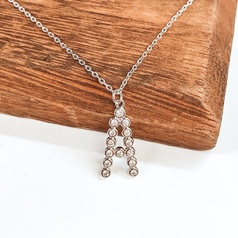 Silver chained necklace with a silver initial "A" pendant that has circle, clear crystals put together to form the initial. This necklace is pictured laying on a brown block on a white background.