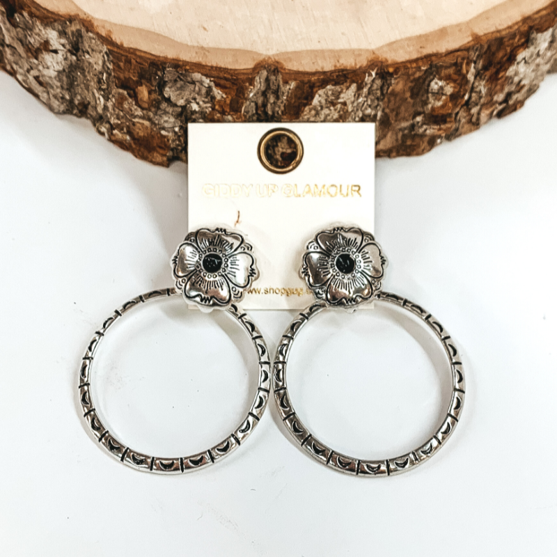Silver, circle post back earrings with flower engraving details and small, black center stone. There is an open, circle pendant with engraving details. These earrings are pictured in front of a piece of wood on a white background.