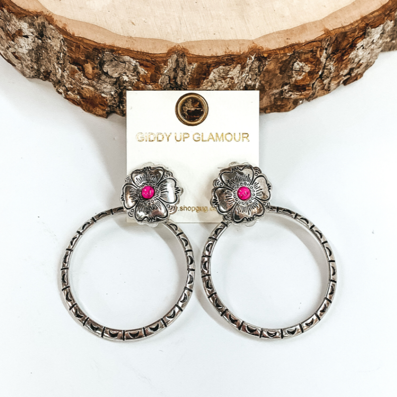 Silver, circle post back earrings with flower engraving details and small, pink center stone. There is an open, circle pendant with engraving details. These earrings are pictured in front of a piece of wood on a white background.