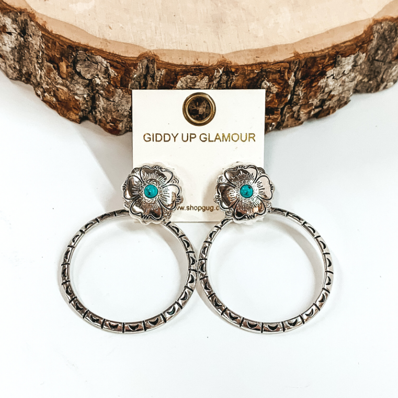 Silver, circle post back earrings with flower engraving details and small, turquoise center stone. There is an open, circle pendant with engraving details. These earrings are pictured in front of a piece of wood on a white background.