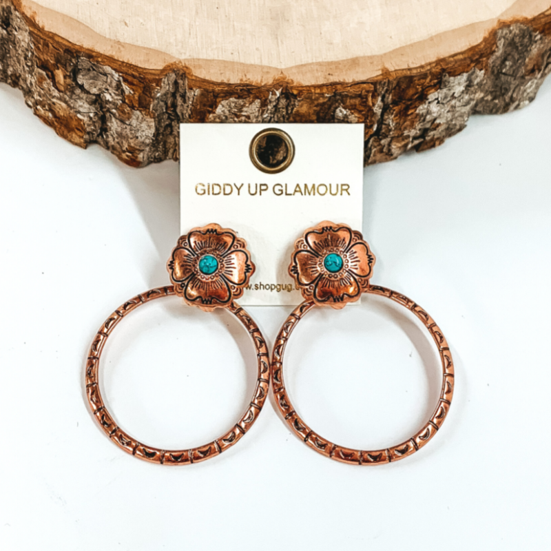 Copper, circle post back earrings with flower engraving details and small, turquoise center stone. There is an open, circle pendant with engraving details. These earrings are pictured in front of a piece of wood on a white background.