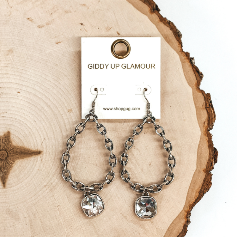 Silver, chained, teardrop dangle earrings with a cushion cut drop clear crystal. These earrings are pictured on a piece of wood on a white background. 