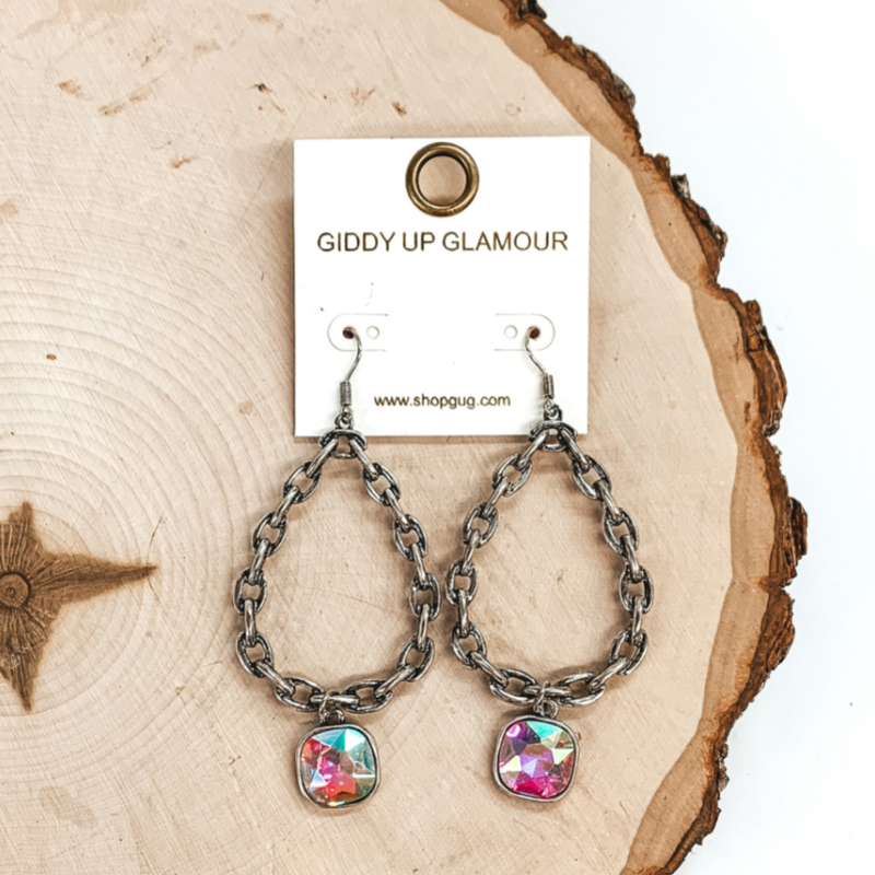 Silver, chained, teardrop dangle earrings with a cushion cut drop AB crystal. These earrings are pictured on a piece of wood on a white background. 