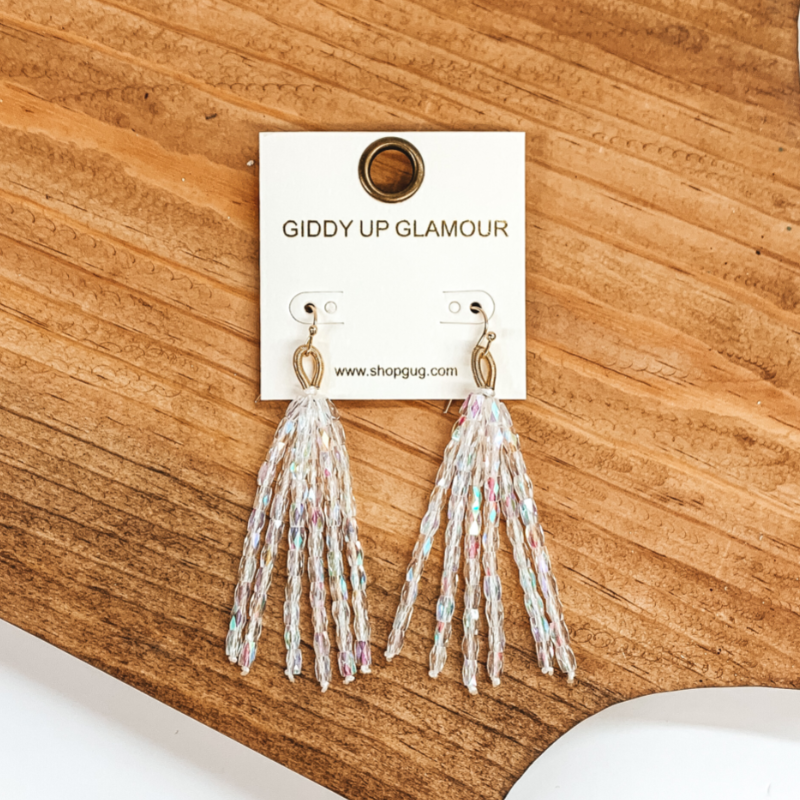 Gold fish hook with upside down gold loop with hanging beaded tassels. The beads are clear crystal beads. These earrings are pictured on a brown block on a white background.  