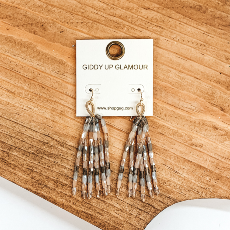 Gold fish hook with upside down gold loop with hanging beaded tassels. The beads are crystal beads in a nude mix that includes grey, charcoal, beige, and ivory. These earrings are pictured on a brown block on a white background. 