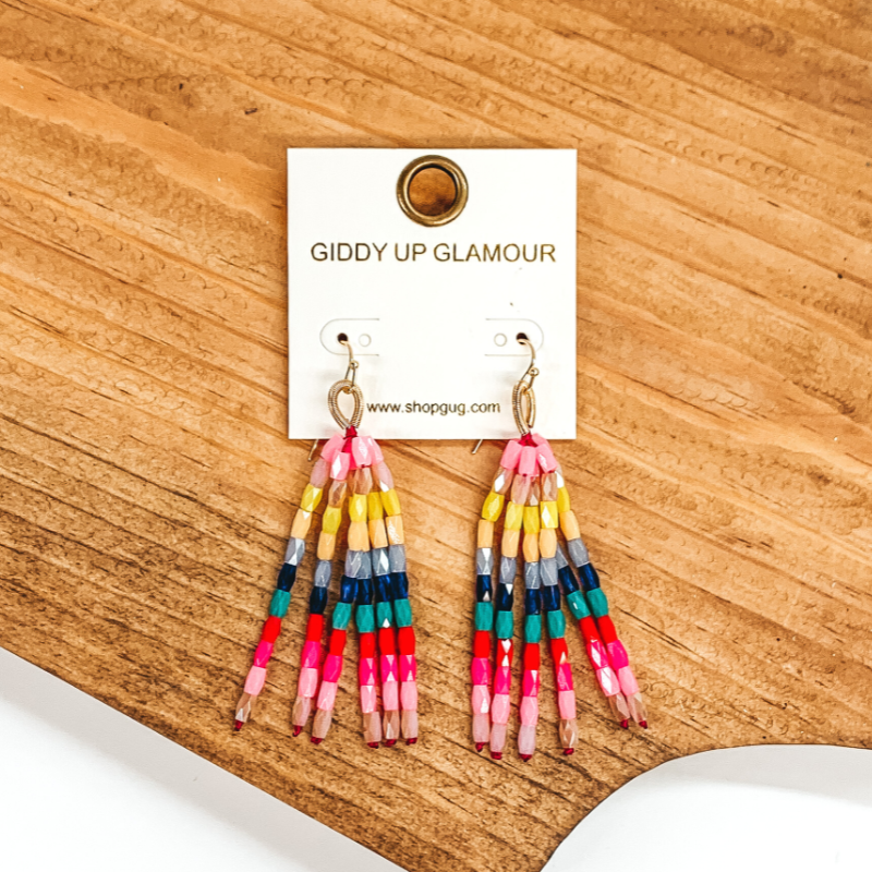 Gold fish hook with upside down gold loop with hanging beaded tassels. The beads are crystal beads in a multicolored mix that includes pink, blue, yellow, teal, and red colors. These earrings are pictured on a brown block on a white background.  