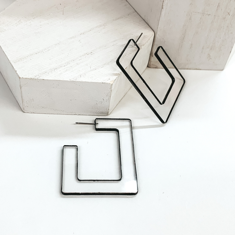 Flat, clear acyrilic square shaped hoop earrings. These earrings have a black trim around the edge. One earring is pictured laying against a white box and the other is laying on the white background.