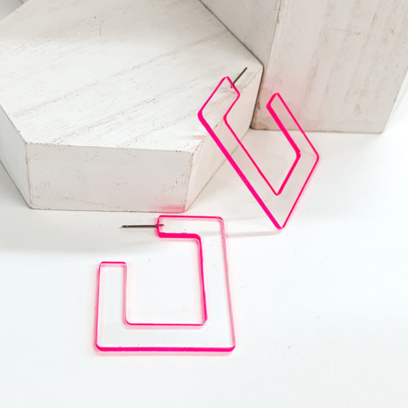 Flat, clear acyrilic square shaped hoop earrings. These earrings have a neon pink trim around the edge. One earring is pictured laying against a white box and the other is laying on the white background. 