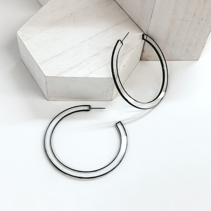 Flat, clear acyrilic circle shaped hoop earrings. These earrings have a black trim around the edge. One earring is pictured laying against a white box and the other is laying on the white background.