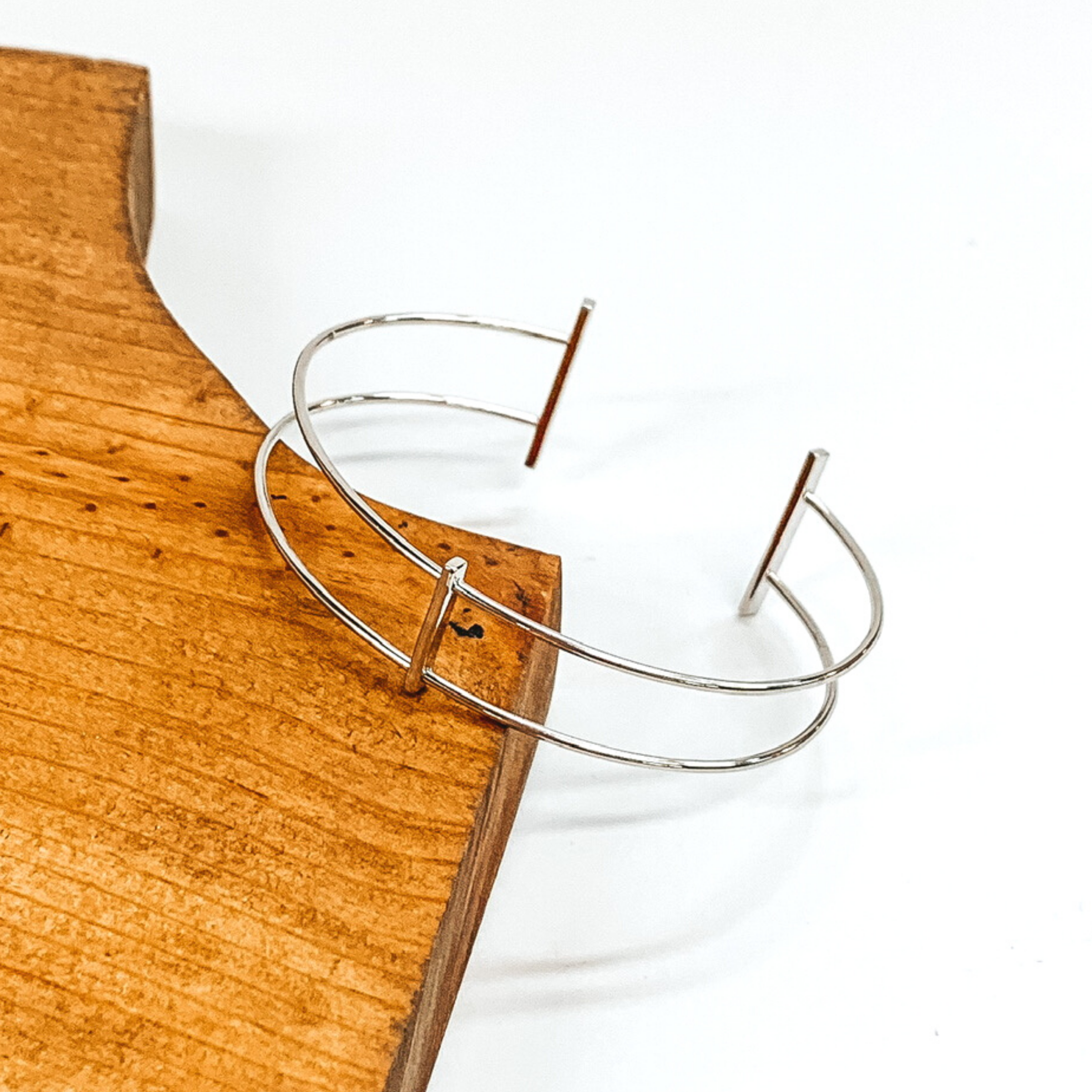 Thin, double wire bangle in silver. The center of the bangle is a plain, thin bar connecting the two wires. The ends have two silver bars. This bracelet is pictured laying on a brown block on a white background. 