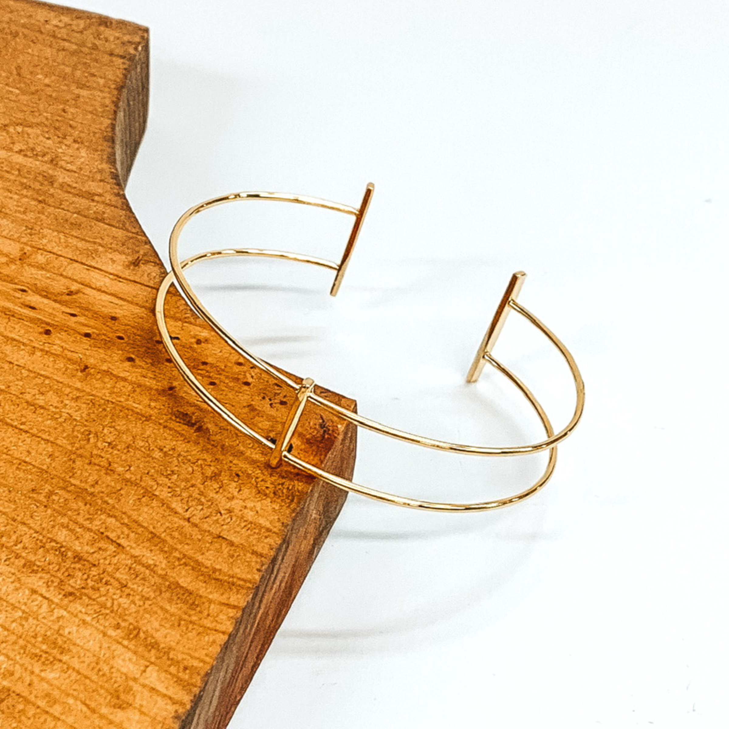 Thin, double wire bangle in gold. The center of the bangle is a plain, thin bar connecting the two wires. The ends have two gold bars. This bracelet is pictured laying on a brown block on a white background.