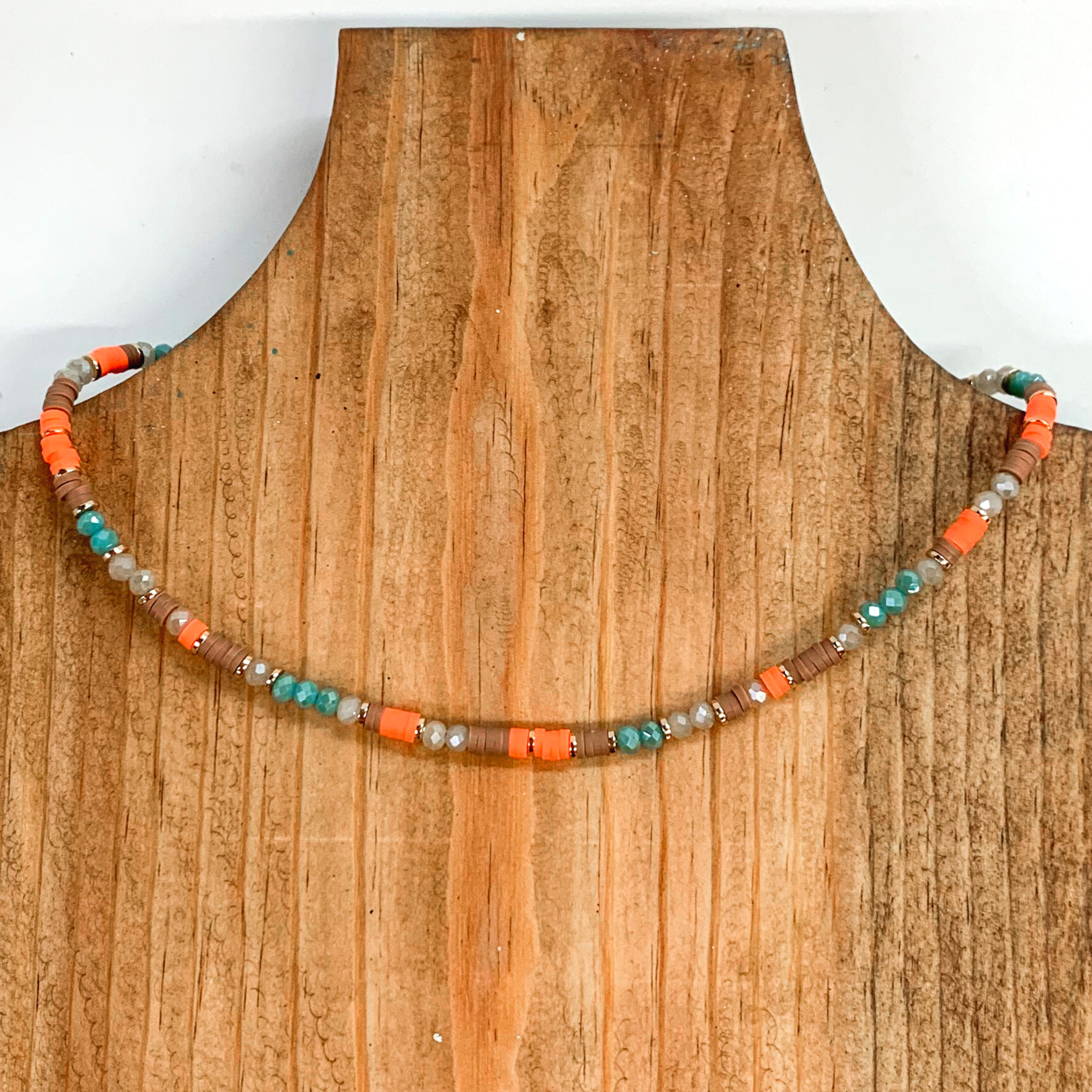 This necklace has segments of crystal beads and rubber beads with gold bead spacers. This necklace includes the crystal beads in the colors teal and a dark beige. The rubber beads come in the colors tan and coral. This necklace is pictured on a laying on a brown necklace holder on a white background.