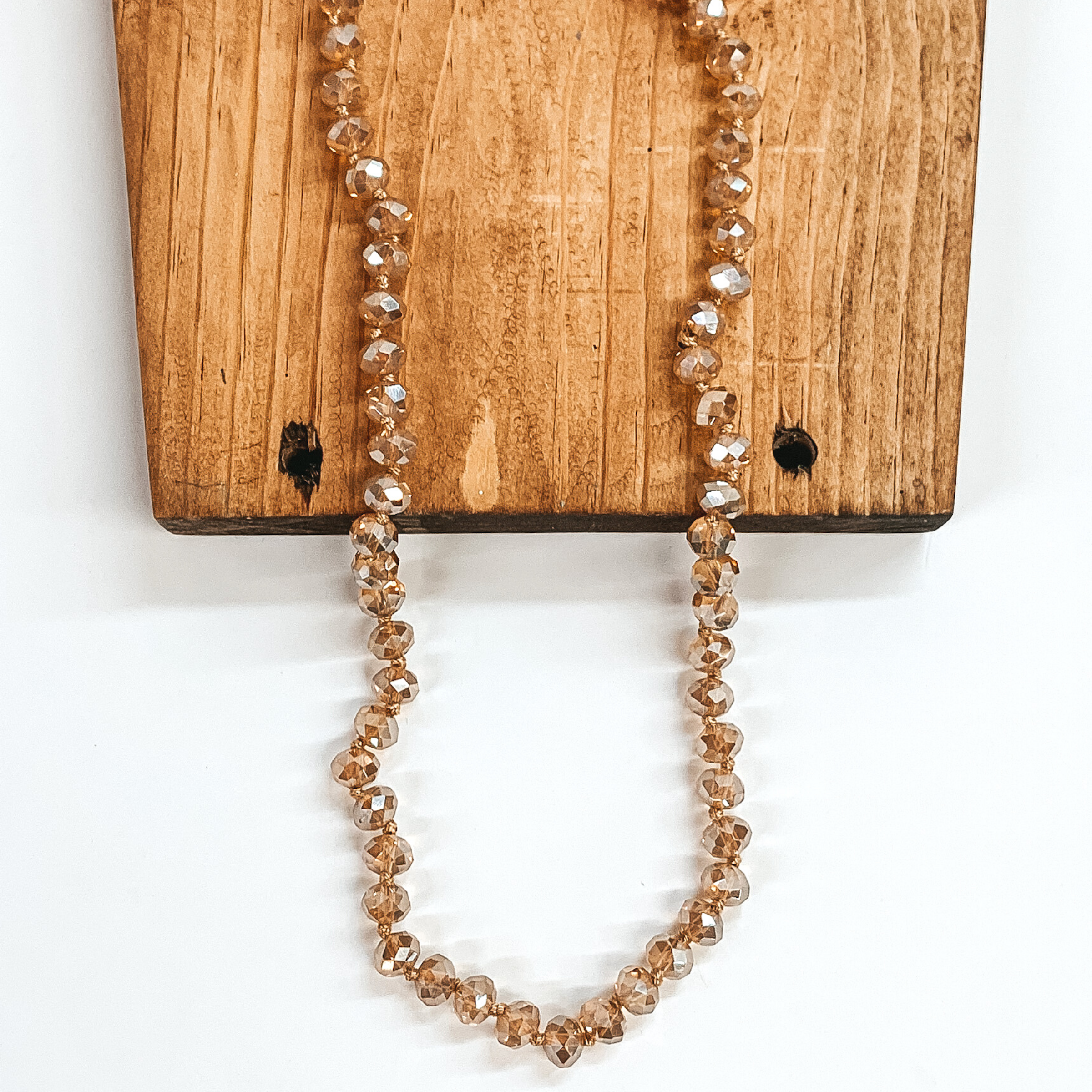 Tan crystal beaded necklace. This necklace is pictured partially laying on a brown block on a white background. 