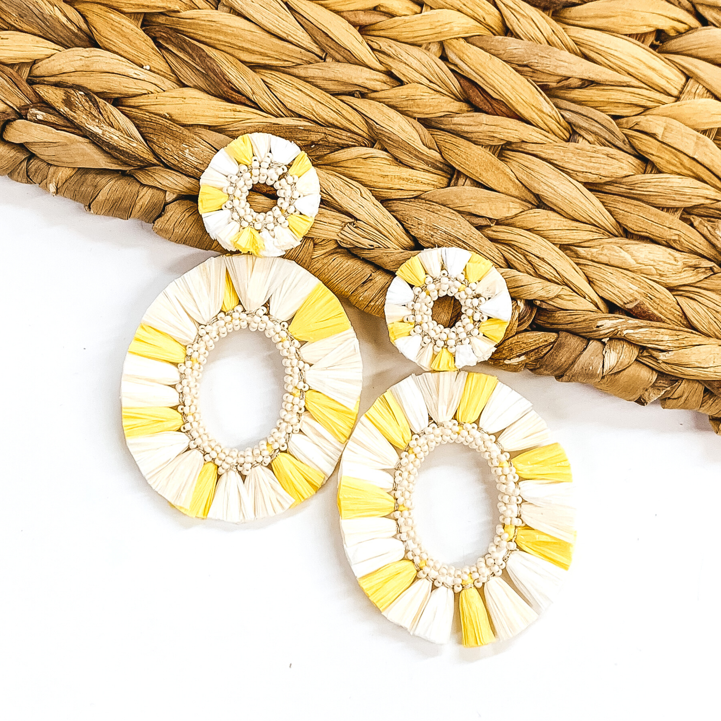 Circle, open beaded earrings with raffia outline. These earrings include an open, circle beaded pendant with raffia outline. The earrings are ivory and yellow colored. These earrings are pictured on a white backround while partially laying on a tan woven material. 