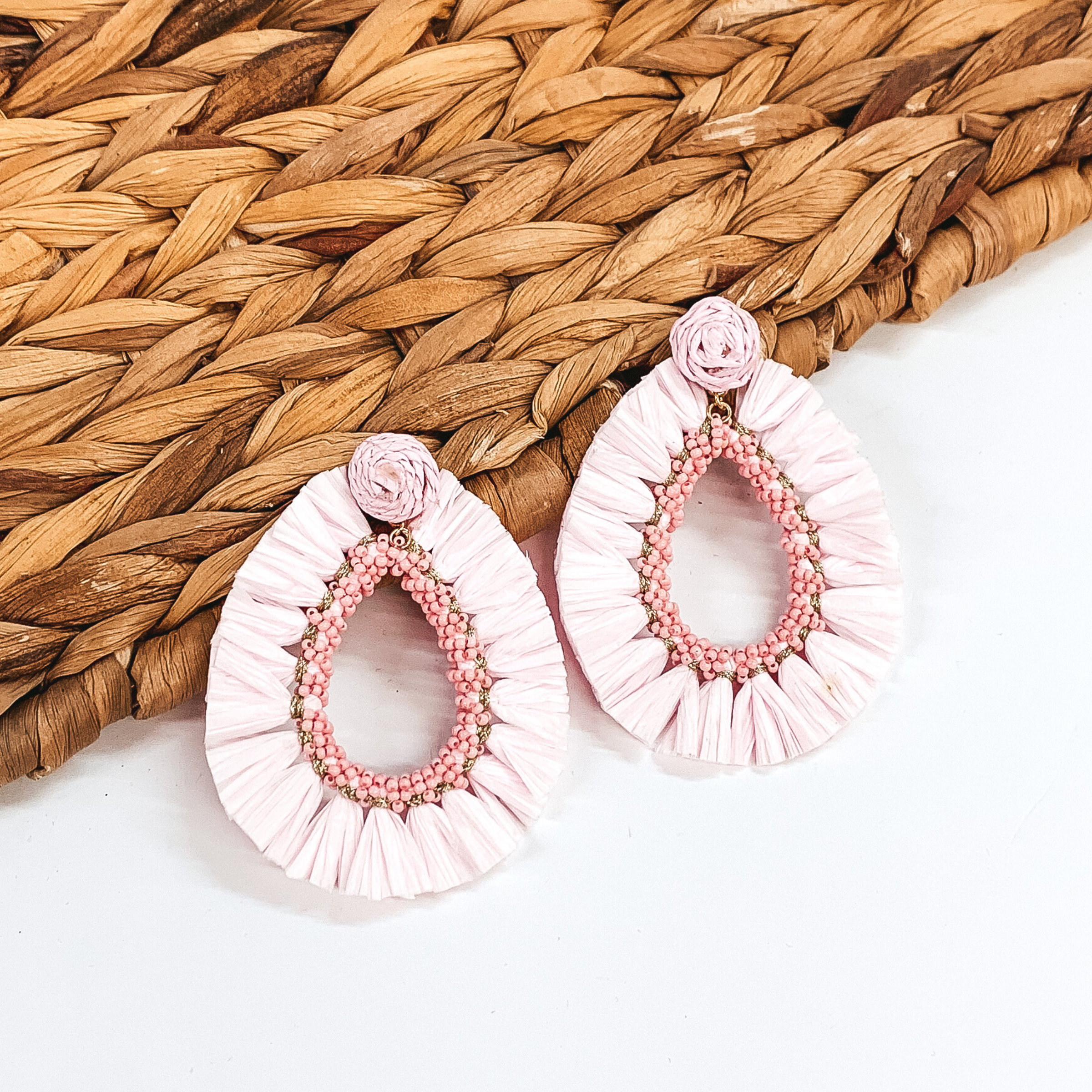 Circle beaded earrings with a hanging open teardrop pendant. The pendant is a beaded teardrop with raffia wrapped around the edges. The earrings are baby pink colored. These earrings are pictured on a white backround while partially laying on a tan woven material. 