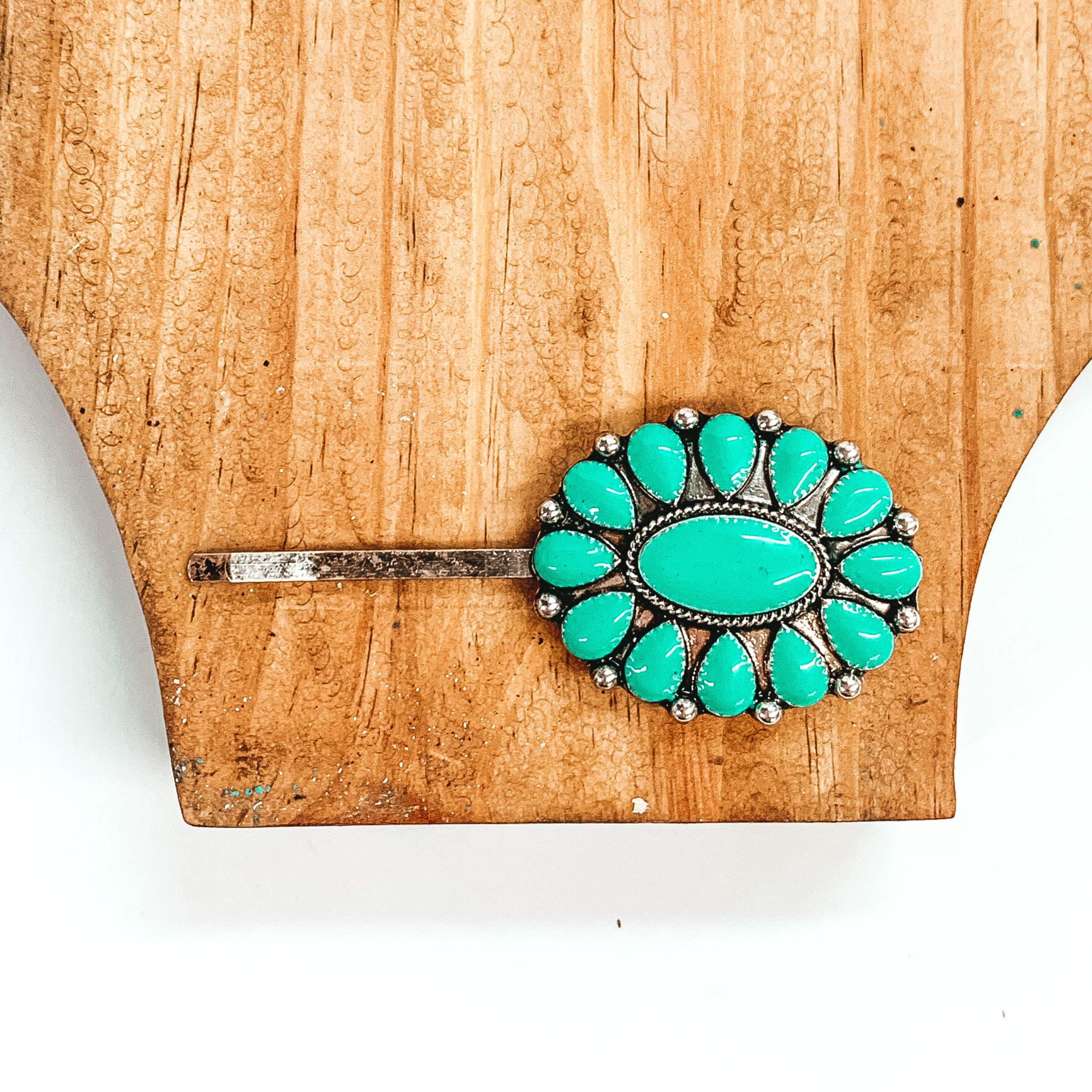 Silver hair pin with a silver oval pendant that has turquoise stones. This hair pin is pictured on a brown block on a white background. 