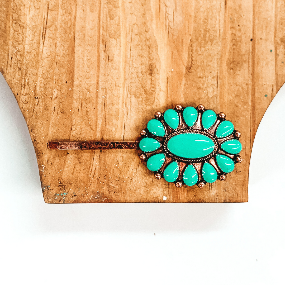 Copper hair pin with a copper oval pendant that has turquoise stones. This hair pin is pictured on a brown block on a white background. 
