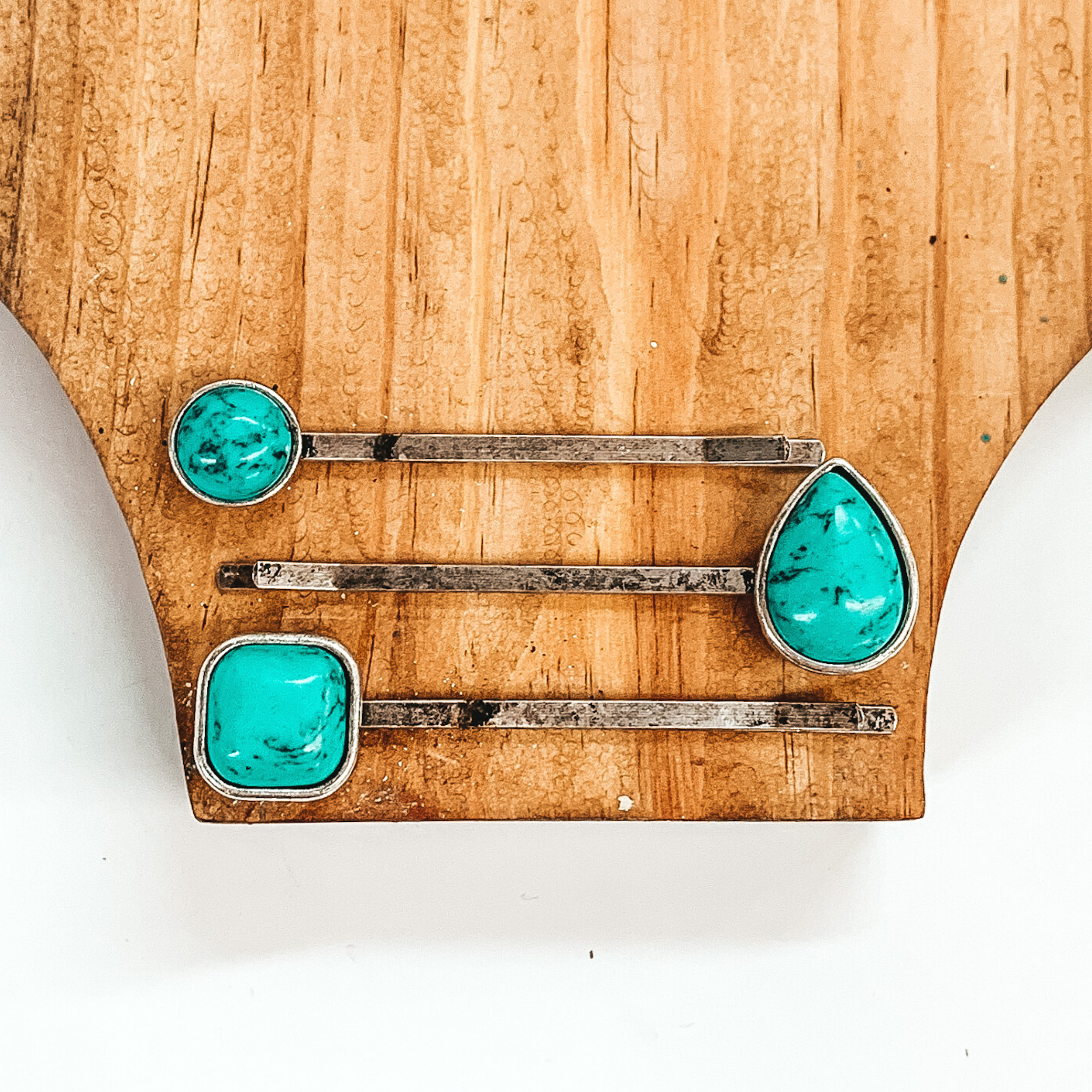 Set of three silver hair pins. Each hair pin has a single turquoise stone in a different shape. These shapes include a circle, teardrop, and a square. These hair pins are pictured on a brown block on a white background. 